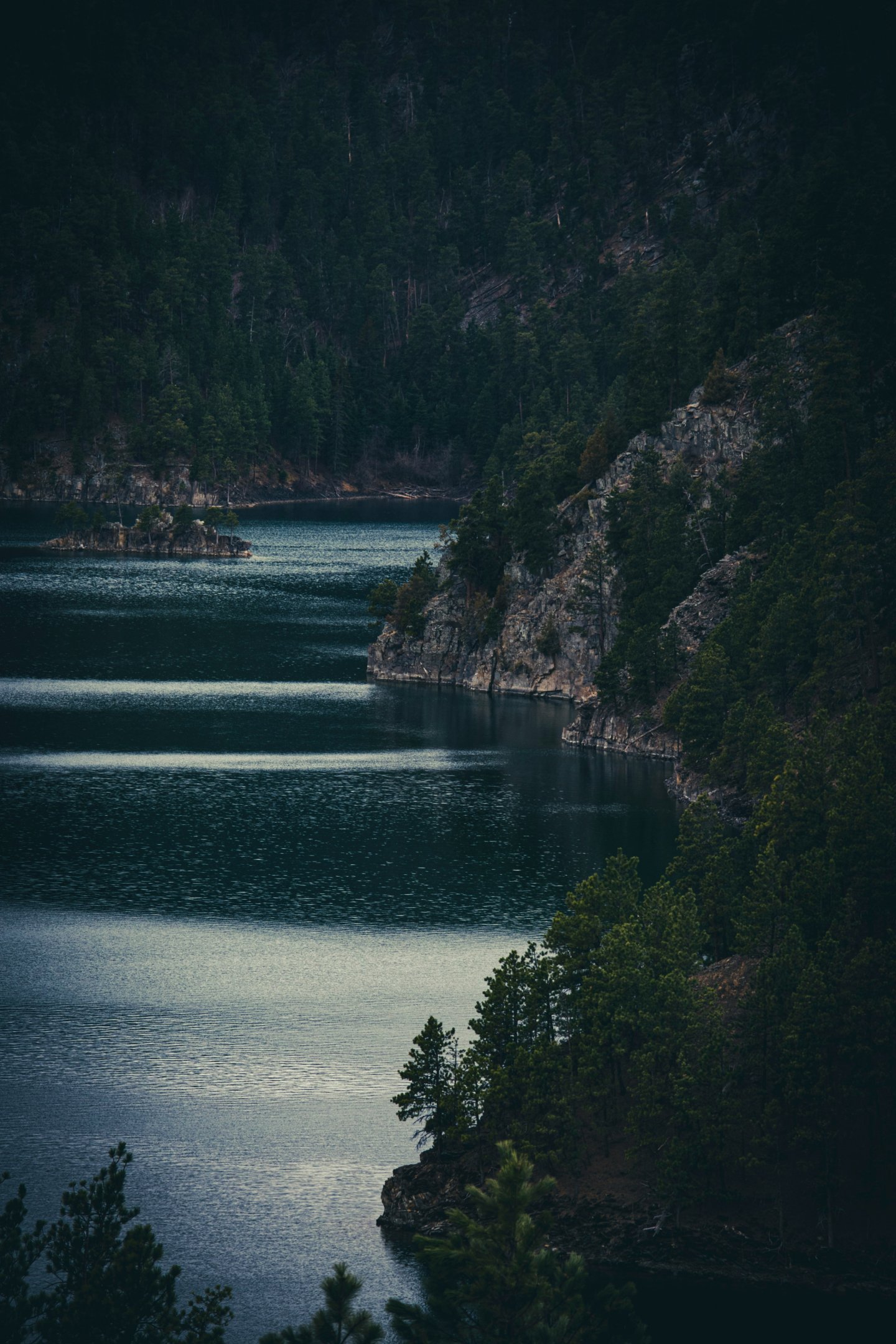 Vue paisible d’un lac de montagne aux eaux sombres, entouré de falaises rocheuses couvertes de pins