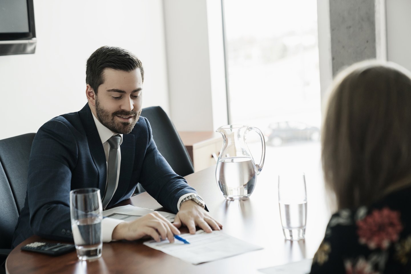 Conseiller financier en complet bleu assis à une table de réunion, souriant en expliquant des documents à une cliente; carafe et verres d’eau sur la table, grande fenêtre en arrière-plan.