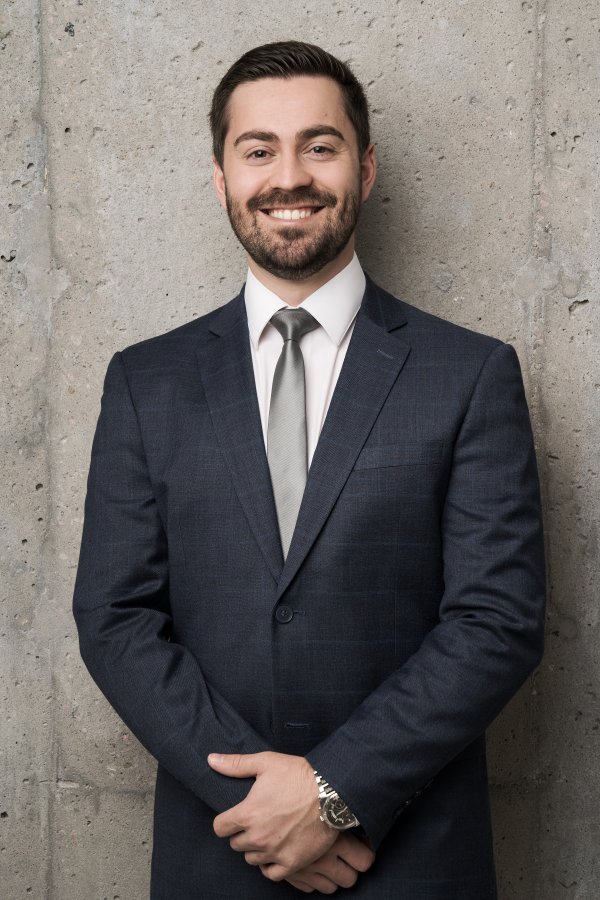 Portrait professionnel d’Antoine St-Pierre, conseiller financier, vêtu d’un complet bleu et d’une cravate gris clair, souriant et debout devant un mur en béton clair.