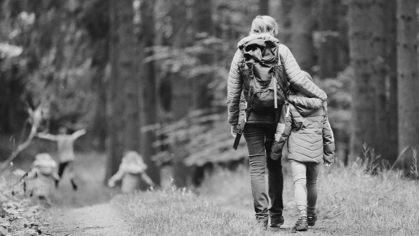 Femme au sac à dos marchant de dos dans un sentier forestier, le bras autour d’une fillette en veste matelassée, tandis que deux autres enfants courent et jouent en arrière-plan.